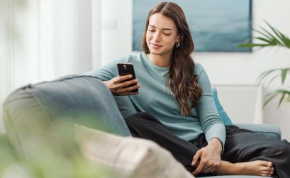 Happy young woman sitting on the couch at home and chatting with her smartphone, she is social networking and using mobile apps