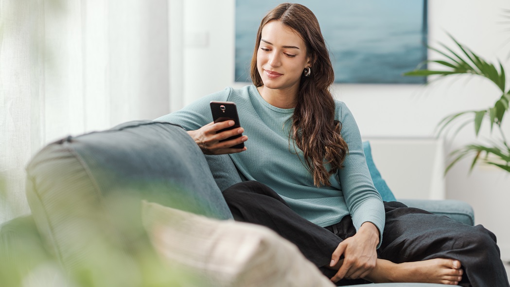 Happy young woman sitting on the couch at home and chatting with her smartphone, she is social networking and using mobile apps